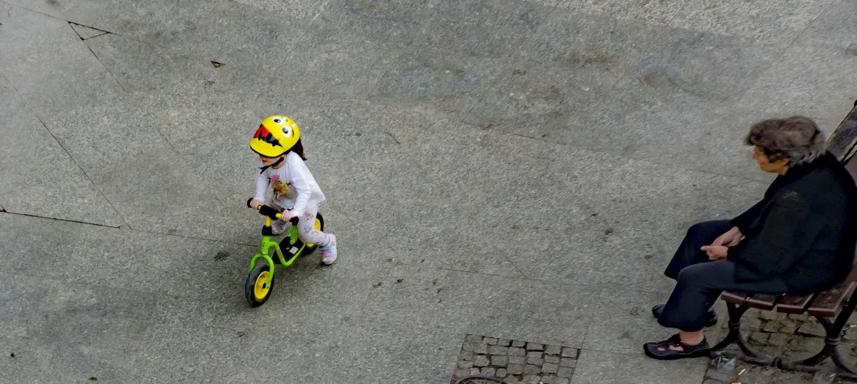 Child on a balance bike wearing a yellow helmet passing an elderly woman on a bench